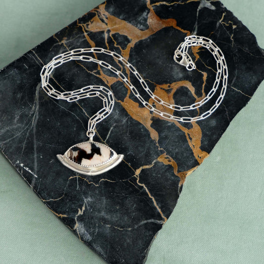 Sterling Silver gemstone Pendant on short paperclip style chain. The pendant features a marquise-cut peanut Wood Cabochon with a striking zigzag pattern in black & white, a serrated sterling silver bezel, and a bamboo style halo mount, cupping the stone. Shown on Black Marble & duck egg velvet background.