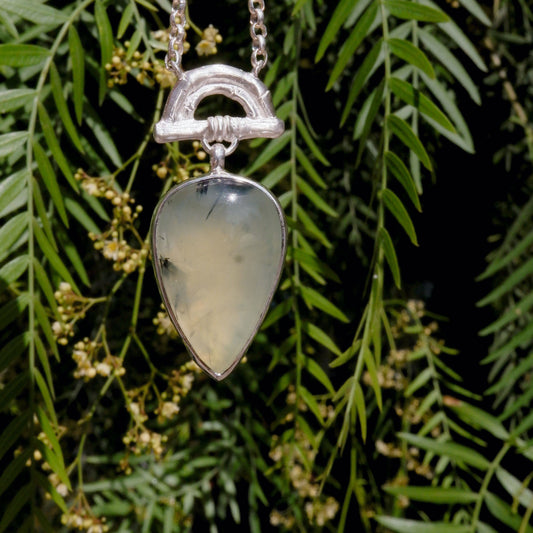 Pendant with Pear-shaped pale green Prehnite Gemstone hanging from silver Bamboo Arch, on fine silver chain. Photographed against green foliage