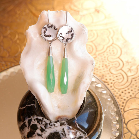 sterling silver earrings featuring mother of pearl discs, engraved with a blackened snake design, and adorned with long green faceted chrysoprase gemstone drop pendants. Shown hanging from an oyster shell in the sunshine.