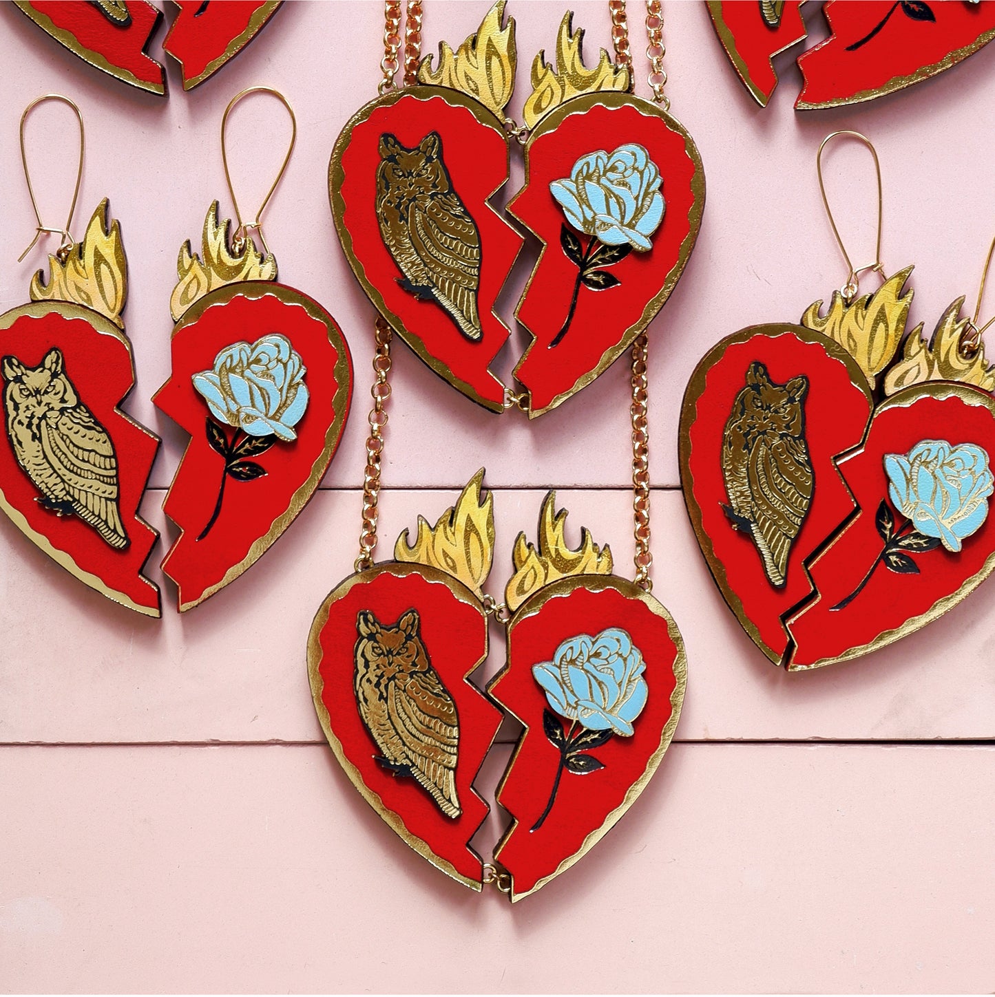 red leather broken heart pendants & earrings, with golden owls, flames & blue roses, laid out flat on pink background