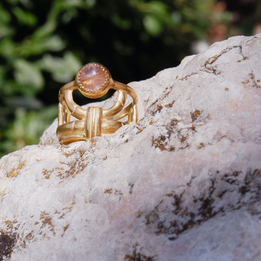 structural open-work ring in a bamboo style, featuring round pale peachy pink Tourmaline cabochon gemstone in a serrated setting. Gold Vermeil. Shown sitting on a rock
