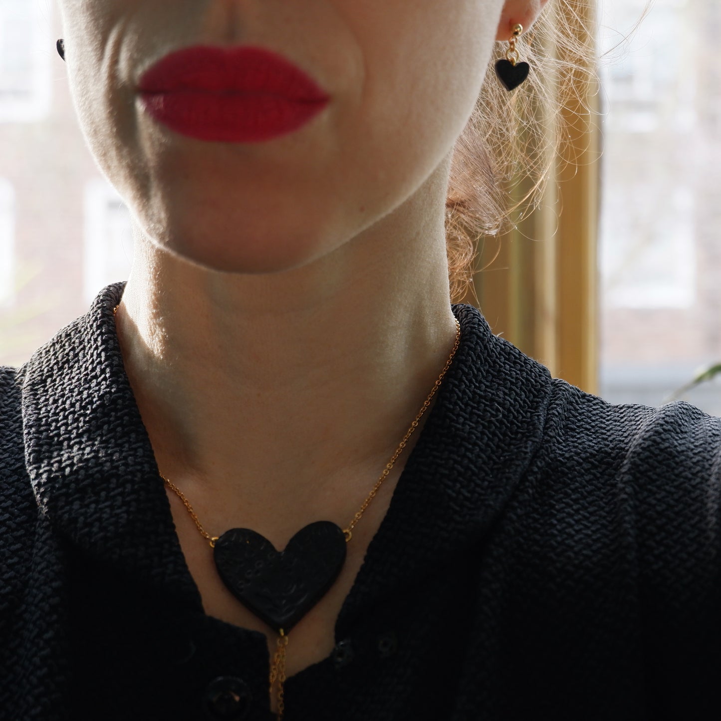 Gold ball stud earrings, with small black, embossed leather heart pendants., with matching large Black Heart pendant necklace, on model in black dress & red lipstick.