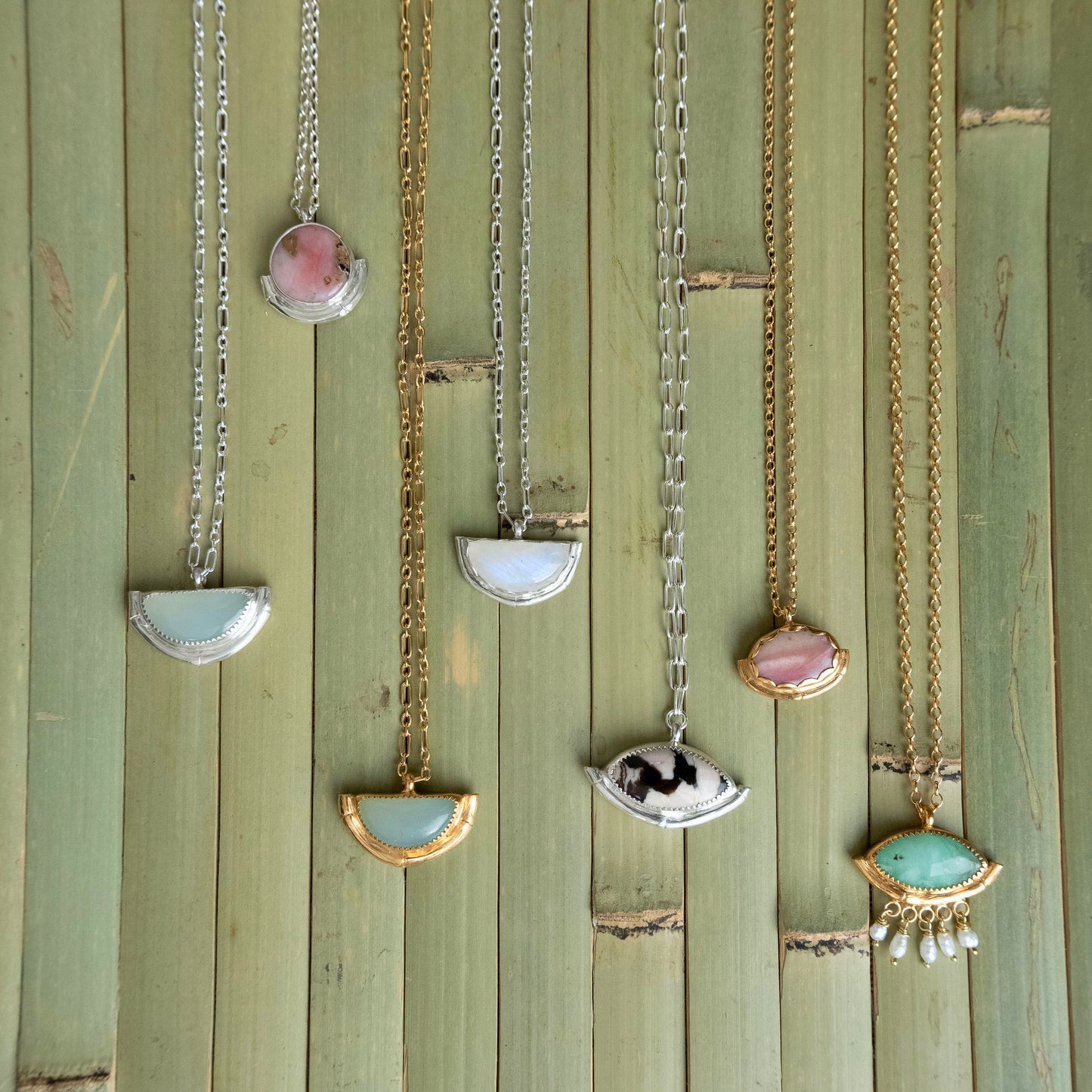 A collection of 7 gemstone pendants in different shapes & colours - each with a bamboo style half frame in Sterling Silver, or Gold vermeil, with a variety of styles of chain. Displayed on a green bamboo panelled background.