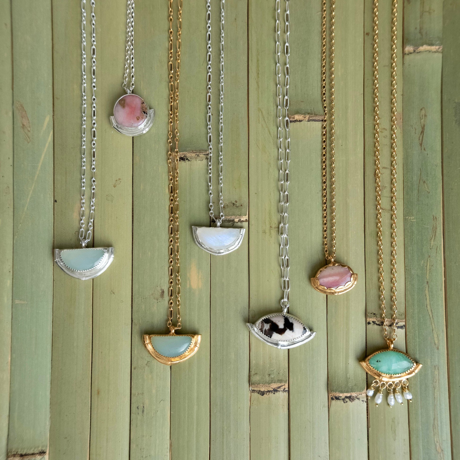 A collection of 7 gemstone pendants in different shapes & colours - each with a bamboo style half frame in Sterling Silver, or Gold vermeil, with a variety of styles of chain. Displayed on a green bamboo panelled background.
