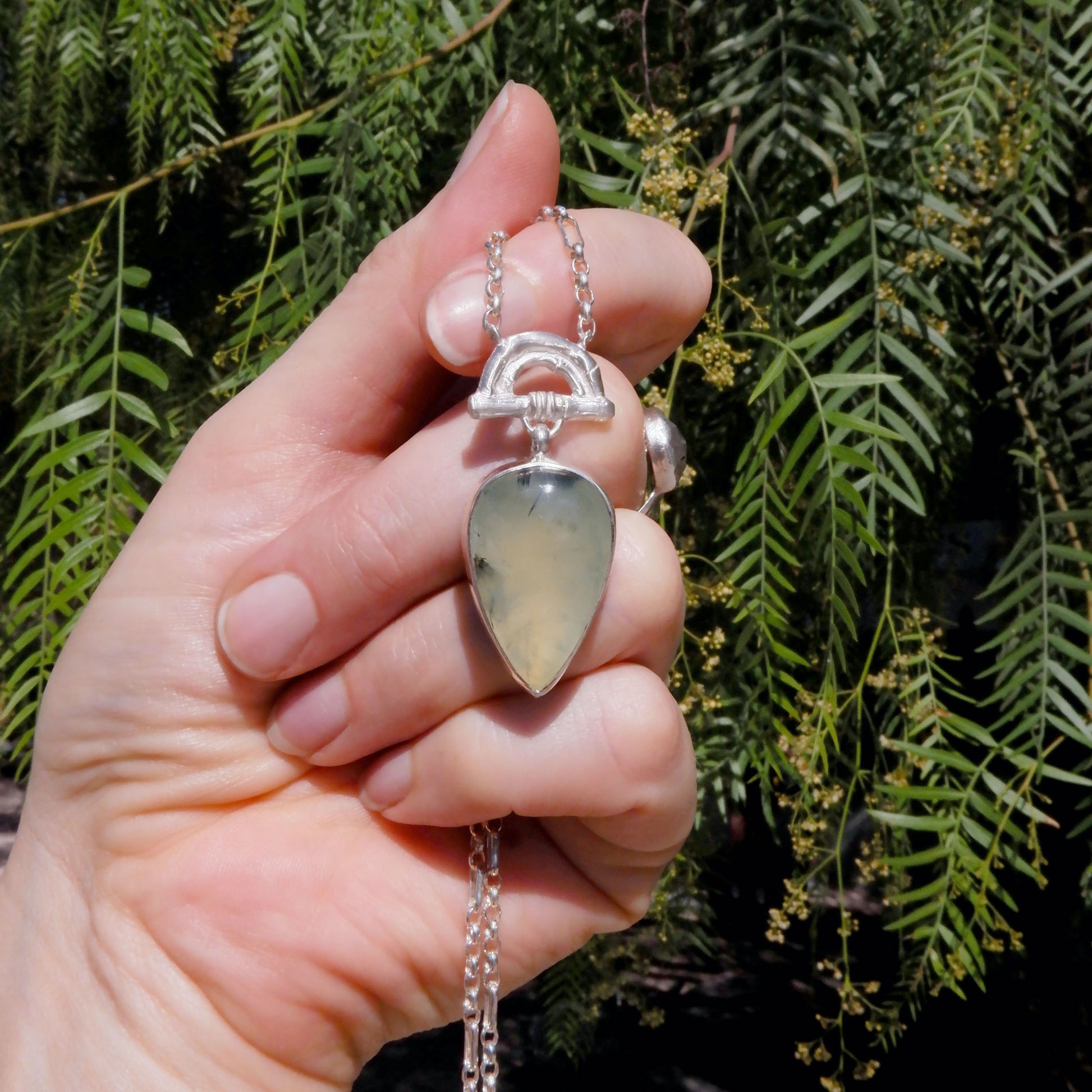 Pendant with  Pear-shaped pale green Prehnite Gemstone hanging from silver Bamboo Arch, on fine silver chain. Photographed in a hand, against green foliage