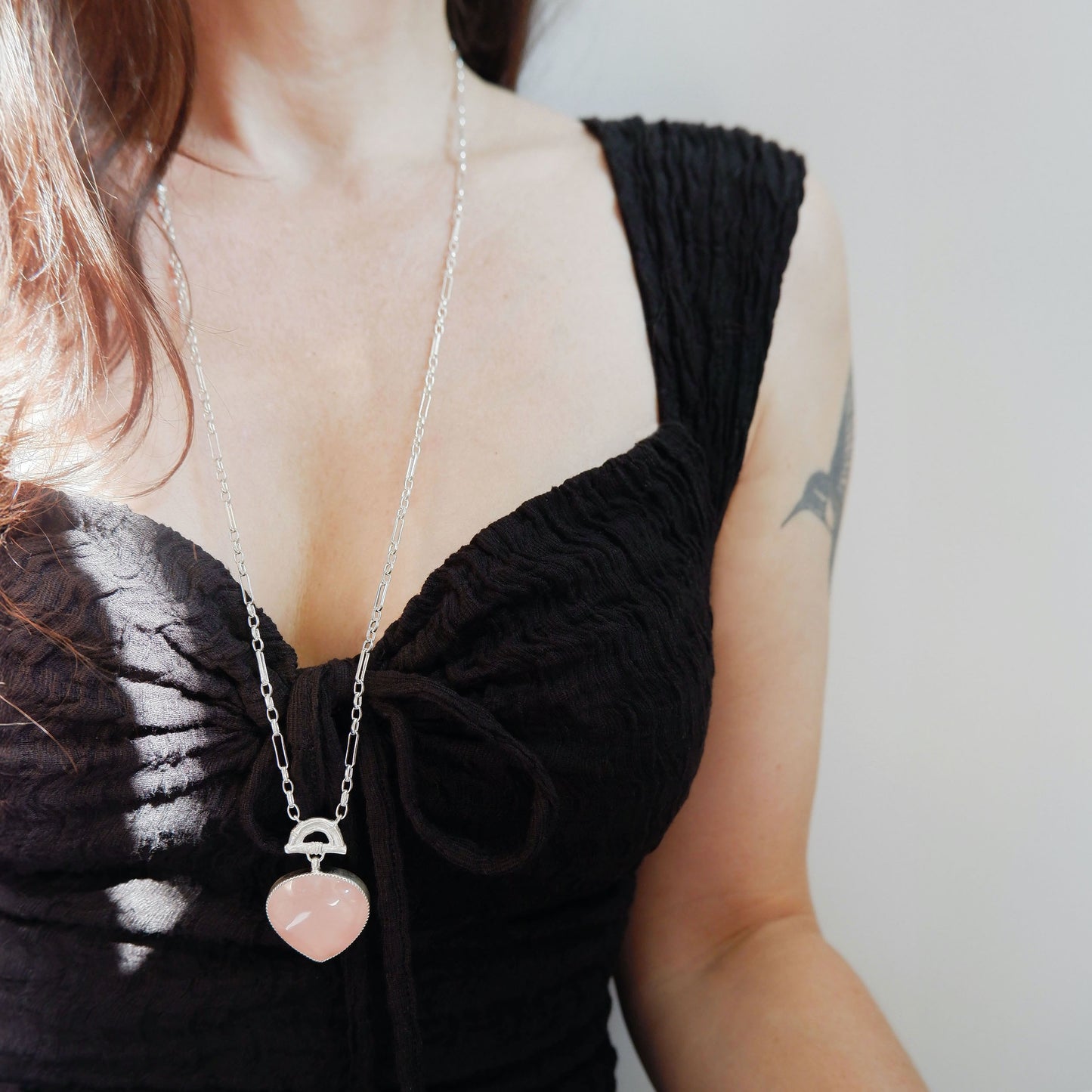 long gold chain necklace with a rose pink quartz heart-shaped pendant with golden bamboo arch detail, on a person in a black dress