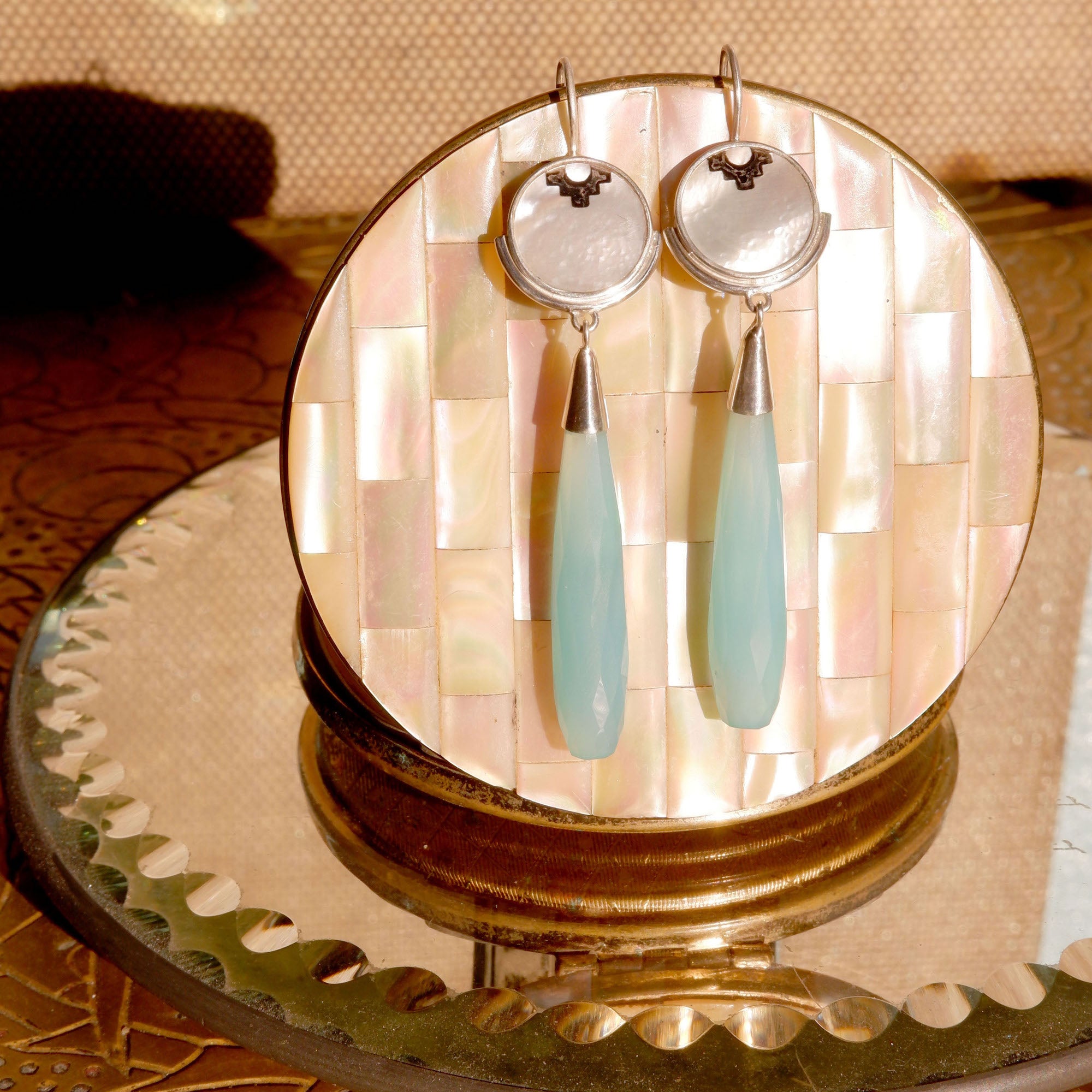 sterling silver earrings featuring mother of pearl discs, engraved with a blackened geometric steps design, and adorned with long blue faceted chalcedony gemstone drop pendants. Shown in still life in warm golden sunshine