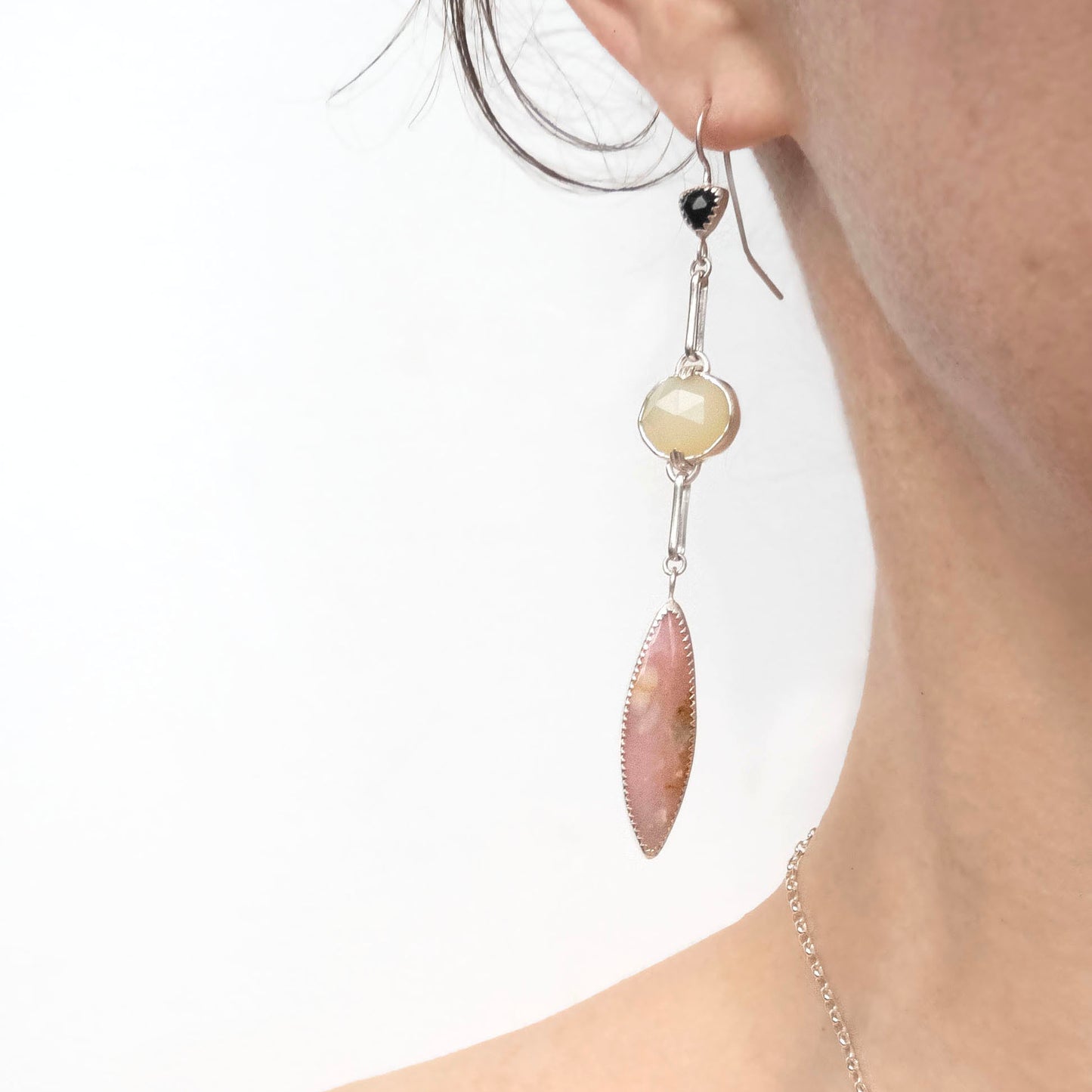 A pair of long drop earrings handcrafted in recycled Sterling Silver, featuring stunning Peruvian Pink Opal Marquise Cabochons, rare faceted butter-yellow Kenyan Opal ovals, and tiny faceted black Onyx Trillions. on person, on White background