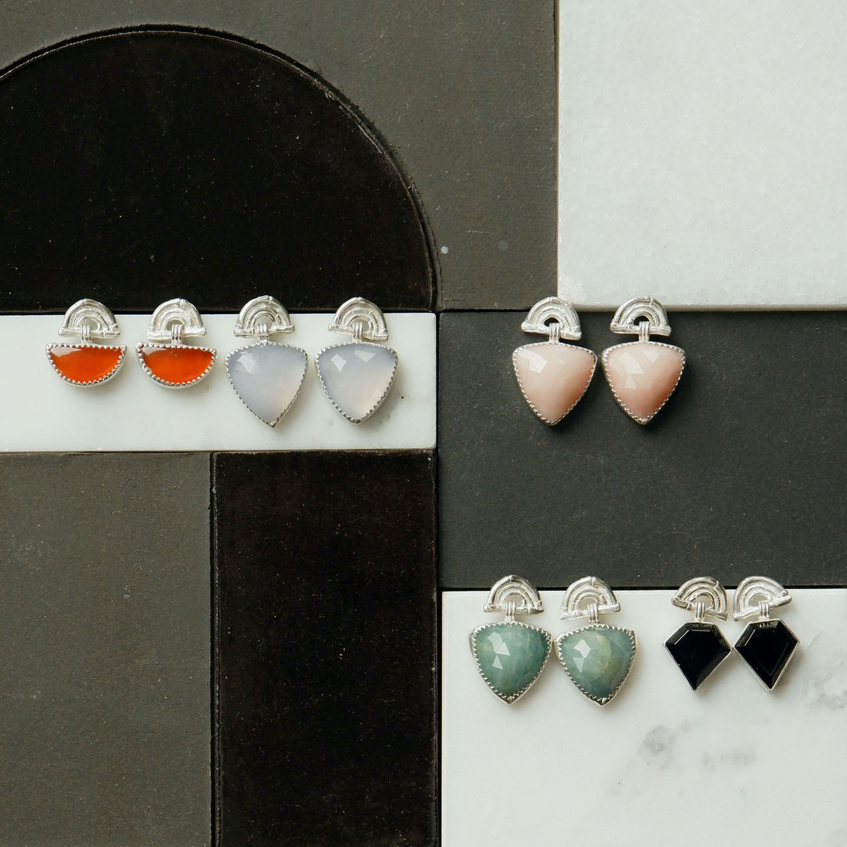 a selection of pairs of sterling silver stud drop earrings , featuring triple arches of bamboo, paired with hanging gemstone charm pendants, set with serrated silver teeth. 
The gemstones shown are half moon shaped deep orange Carnelian, Trillion shaped Lavender Chalcedony, Pink Opal & Aquamarine, and Black Onyx Diamonds.
Shown on black & white tiled background.