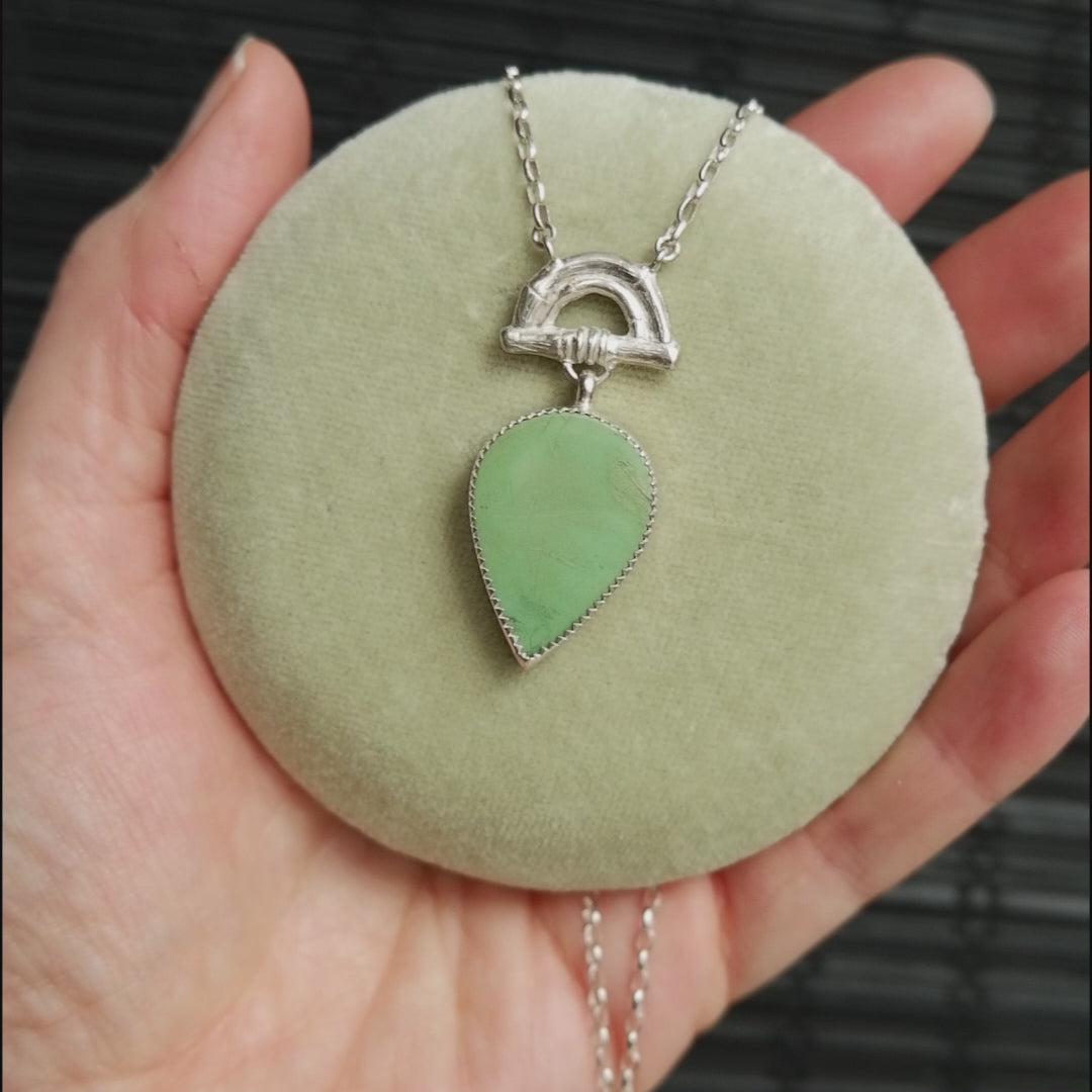 video showing Pendant with green turquoise pear shaped gemstone hanging from silver Bamboo Arch, and silver chain.  shown on pale green velvet