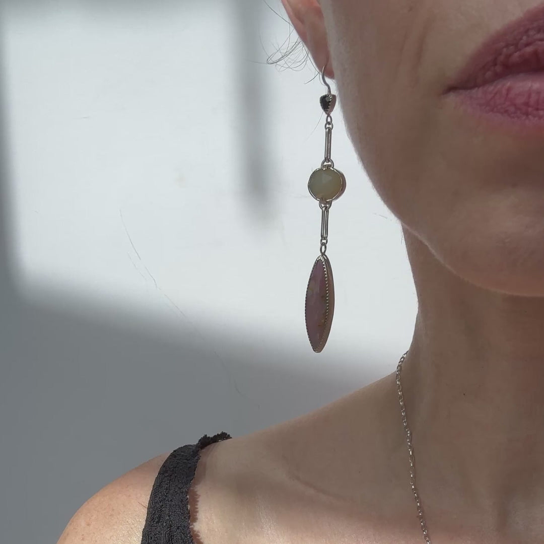 A pair of long drop earrings handcrafted in recycled Sterling Silver, featuring stunning Peruvian Pink Opal Marquise Cabochons, rare faceted butter-yellow Kenyan Opal ovals, and tiny faceted black Onyx Trillions. shown on model. White background