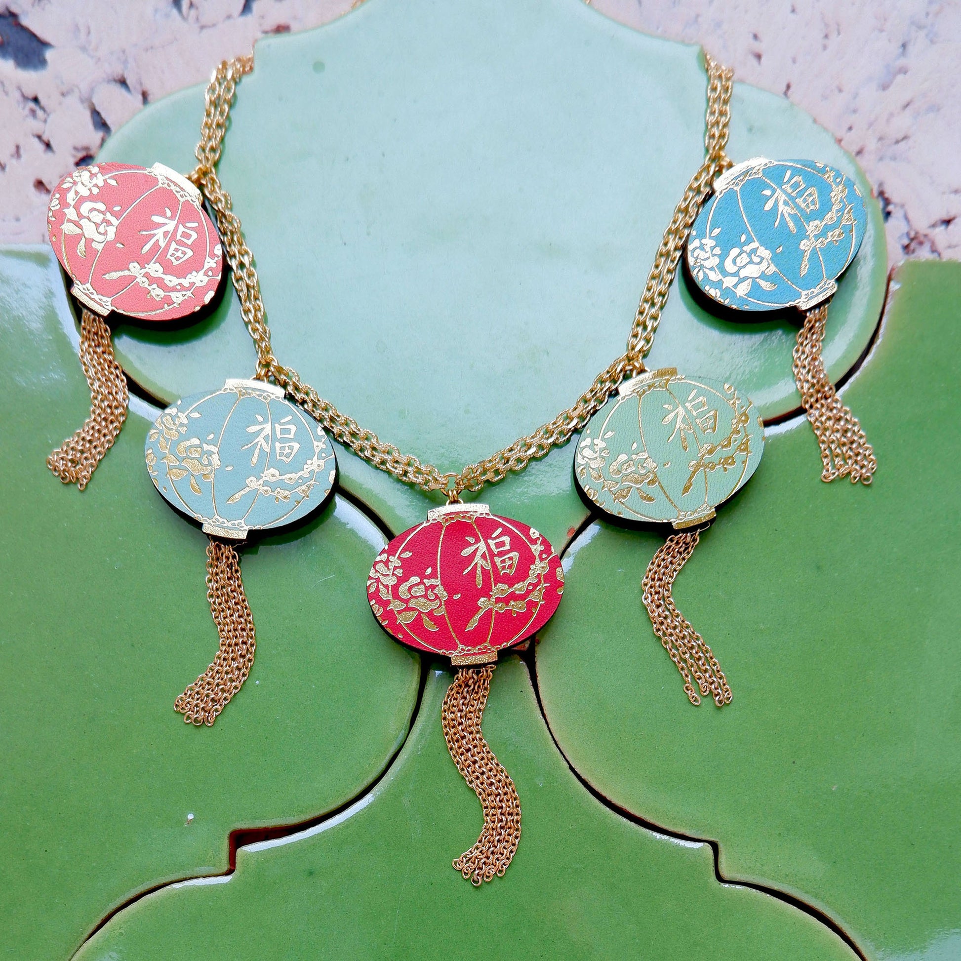 gold chain necklace, with 5 multi coloured leather lantern pendants, or charms. Each one, pink, aqua, scarlet red, light olive green and electric blue, with a gold chain tassel. On green tile background